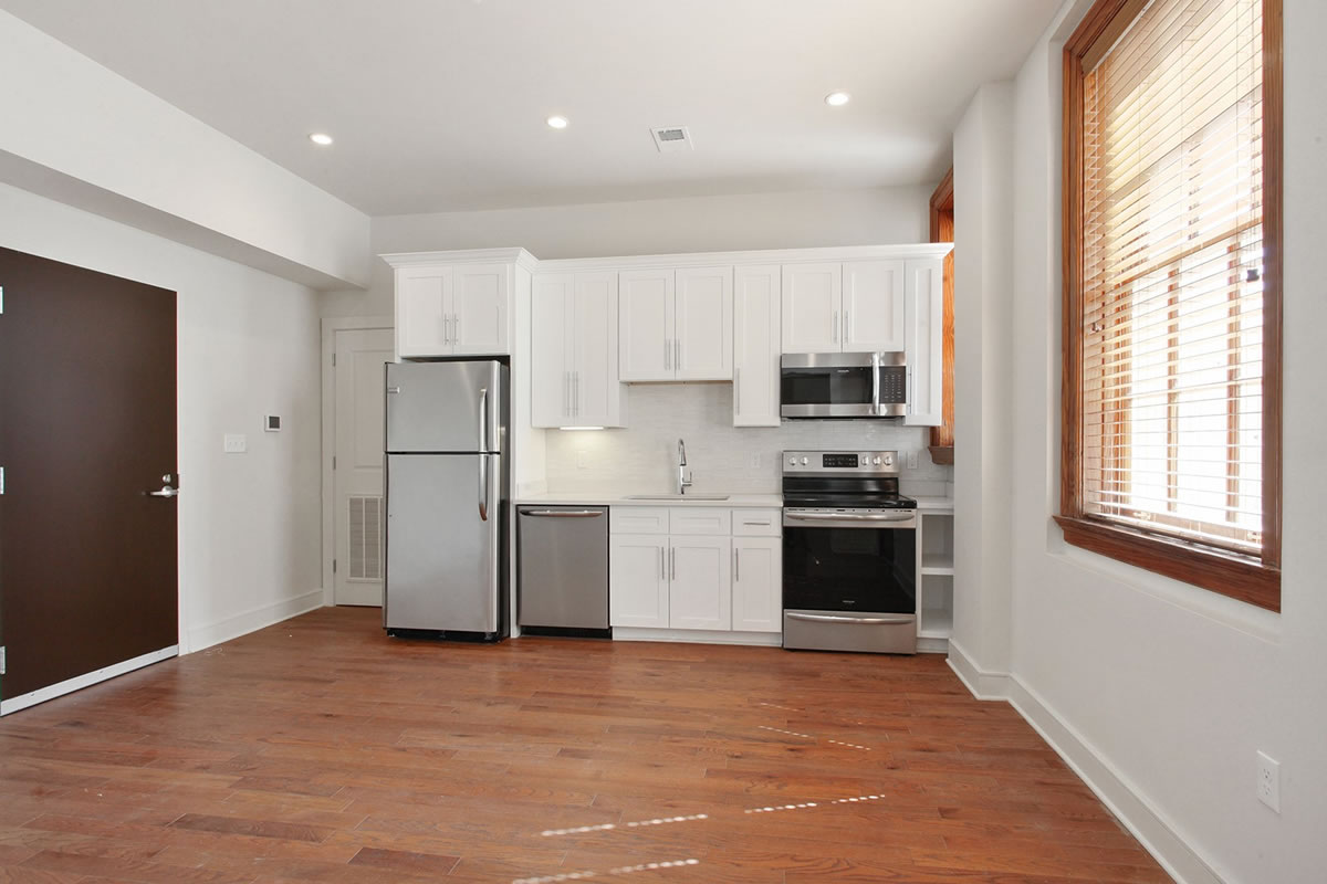 Townhomes For Rent In New Orleans, LA - The Academy - Kitchen With Wood-Style Flooring, High Ceiling, Stainless Steel Appliances, Quartz Countertops, Custom White Cabinets, Built-In Microwave, And A Window With Blinds