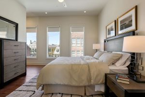 Luxury Apartments For Rent In New Orleans - The Academy - Spacious Bedroom With Wood-Style Flooring, Area Rug, Large Bed, Nightstands With Lamps, Dresser, Mirror, High Ceiling, Ceiling Fan, And Large Windows