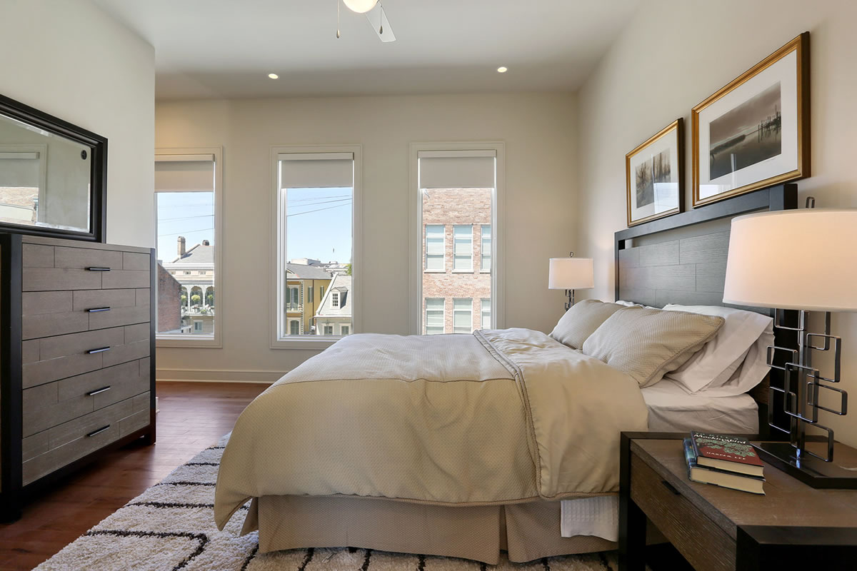 Luxury Apartments For Rent In New Orleans - The Academy - Spacious Bedroom With Wood-Style Flooring, Area Rug, Large Bed, Nightstands With Lamps, Dresser, Mirror, High Ceiling, Ceiling Fan, And Large Windows