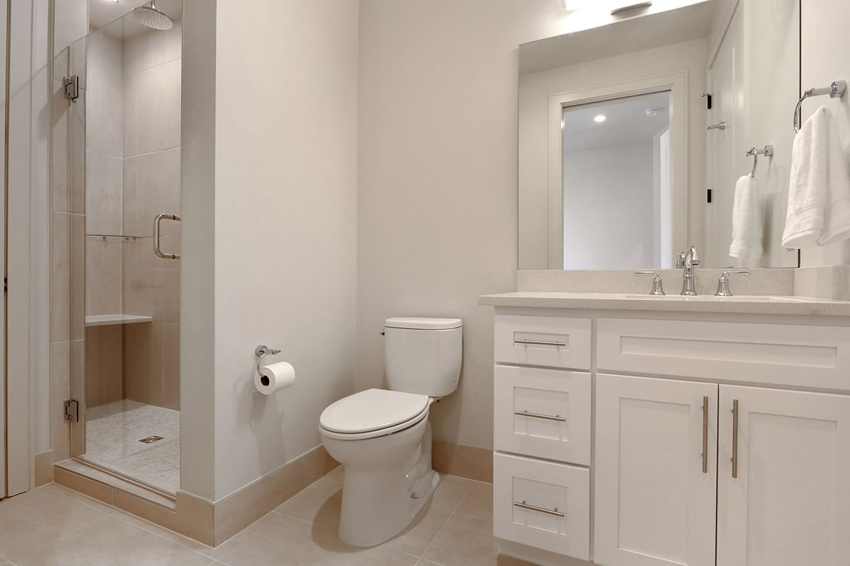New Orleans, LA, Apartments - The Academy - Bathroom With Tiled Flooring, Vanity Mirror, White Cabinets, Toilet, And A Walk-In Shower With Clear Glass Door
